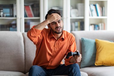 Sad handsome young arab guy in smart casual playing video games alone at home at weekend, man holding joystick, sitting on couch, touching his forehead, lost in game, copy space