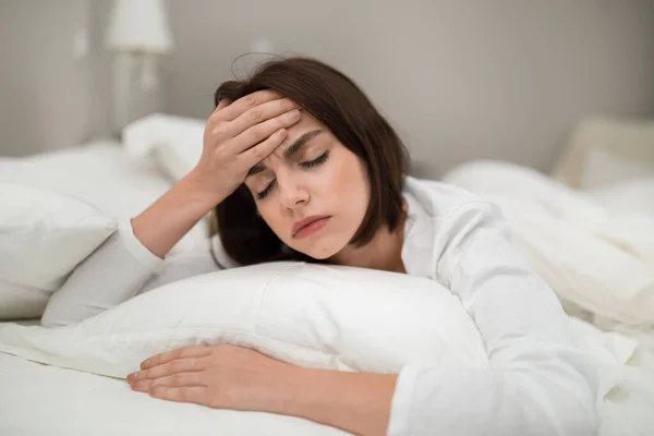 Upset young brunette woman lying in bed at home, touching head and hugging pillow, tired lady wake up with headache in the morning, suffering from chronic migraine, copy space