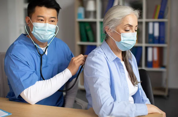 Serious adult korean man doctor in protective mask listens with stethoscope to breathing of old european lady patient in clinic interior. Medical examination, health care, treatment with therapist
