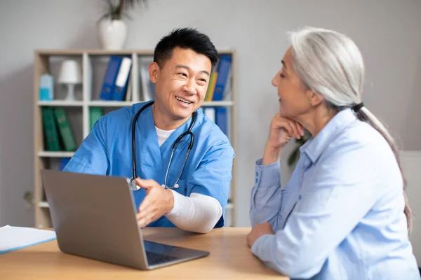 Happy glad handsome middle aged korean doctor shows laptop to old female caucasian patient in clinic office interior. Recommendation and advice from therapist, health care with device and professional