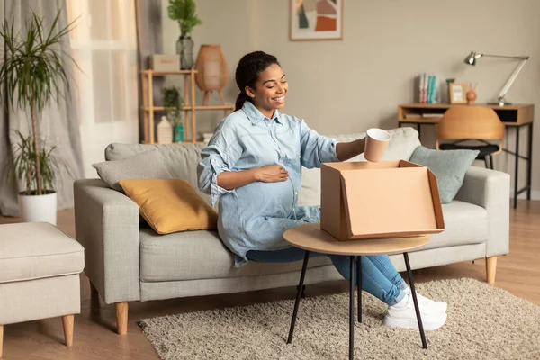 Happy young pregnant black lady unpacking box and holding cup, touching belly and smiling, sitting on sofa at home, free space, full length. Pregnancy lifestyle and delivery service