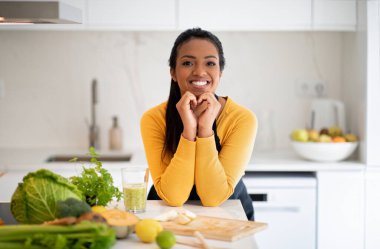 Happy young african american lady make heart sign with hands at table with organic vegetables, fresh smoothie in kitchen interior. Vegan food, cook healthy eat at home, body care and proper nutrilon