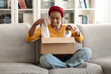 Amazed excited happy young black woman in casual sitting on couch at home, female shopaholic unpacking webstore delivery box parcel, checking her new white shoes sneakers, copy space