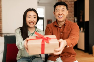 Happy asian couple stretching wrapped gift box to camera and smiling, sitting on couch at home. Excited spouses congratulating with birthday or holiday