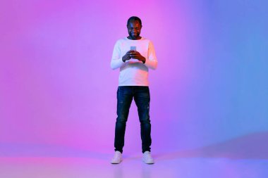 Happy smiling handsome middle aged african american man in casual posing on neon light background, using phone, checking newest mobile app, surfing on Internet, full length studio shot, copy space
