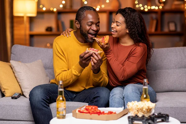 Happy pretty young african american woman feeding her boyfriend while spending romantic evening together, black couple sitting on couch, eating dinner pizza, drinking beer, having fun, home interior