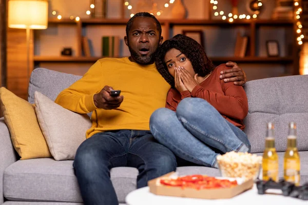 Frightend loving black couple watching horror movie or thriller on TV together at home, african american man and woman sitting on couch, embracing, closing eyes, copy space. Domestic entertainment