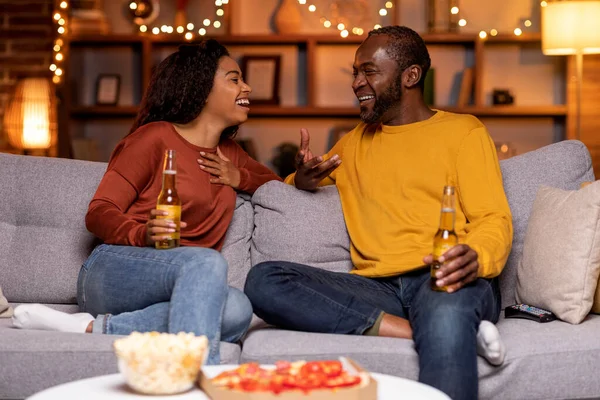 Black couple in love having date at home, happy cheerful african american young woman and middle aged man sitting on couch, drinking beer, eating pizza, having conversation, laughing, copy space