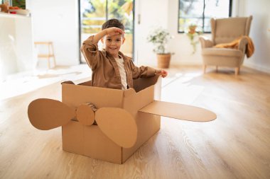 Smiling little european boy in cardboard box helicopter looks at distance in light room interior. Upbringing and childhood, fun and entertainment, art and creative at home during covid-19 pandemic