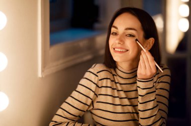 Happy cheerful beautiful brunette young lady in casual sitting in front of mirror, aplying makeup at home, holding visage brush and using eyeshadows or highlighter, smiling, copy space