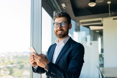 Business and telecommuting concept. Portrait of happy middle aged man using smartphone, texting with client while standing near window in office, free space