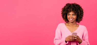 Cheerful millennial african american curly lady with braces in casual typing on phone looks at copy space isolated on pink background, panorama, studio. Blog and social networks, app, ad and offer