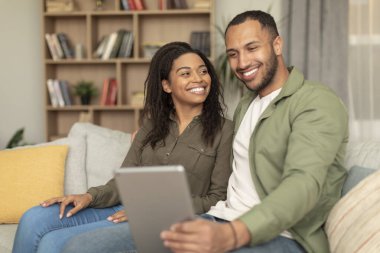 Happy black lovers sitting on couch and using digital tablet, having video call or surfing internet, using newest mobile application, home interior, copy space