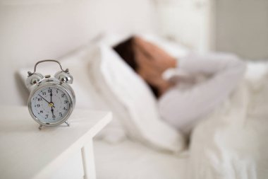 Selective focus on silver mechanical alarm clock on bedside table over blurred sleeping lady, unhappy brunette woman in white pajamas lying in bed in white bedroom at home, covering her face