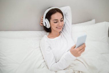 Relaxed cheerful pretty young woman in white cotton pajamas sitting in bed with smartphone and wireless headphones, looking at gadget screen and smiling, watching movie or TV show online, copy space