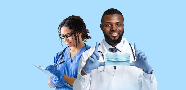 Positive smiling black doctor therapist and female nurse working on blue background, clinician showing protective face mask while female medical worker taking notes at clipboard, panorama