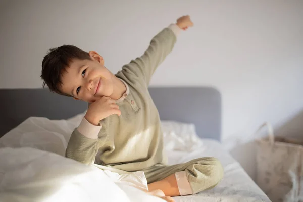 Cheerful energy cute little boy sits on bed, woke up and raises his hand up, stretching body in bedroom interior, free space. Health care, sleep, upbringing and childhood, rest and relax at morning