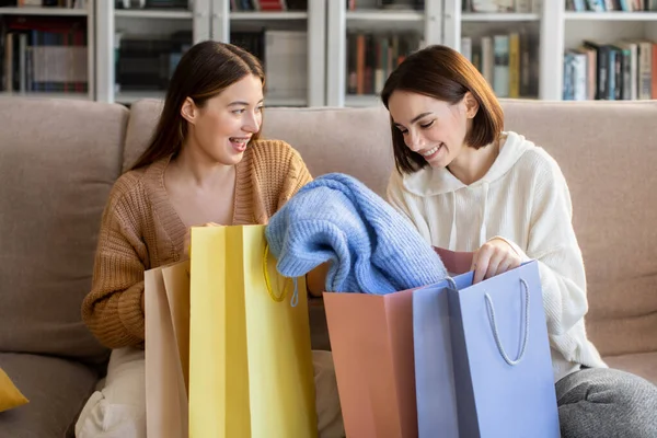 Laughing european millennial ladies shopaholic in sweaters open bags from store, enjoy new clothes in leisure time in comfort living room interior. Relationships, shopping together and sale at home