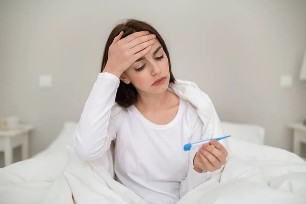 不幸な病気若いブルネットの女性がベッドに座って暖かいDuvet 額に触れ 電子温度計を保持します 発熱に苦しみながら風邪やインフルエンザを持っています — ストック写真