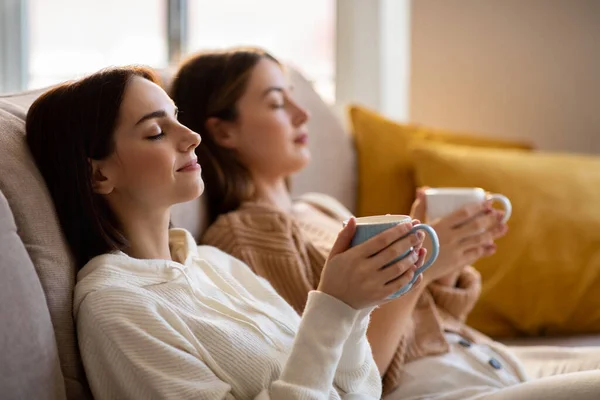 Satisfied european millennial ladies with closed eyes in sweaters with cups of coffee sit on sofa enjoy rest, relax, calm and free time in living room interior. Hot drink break, think and dreams