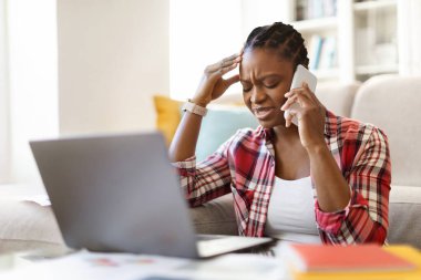 Professional burnout. Depressed upset african female sit on floor by laptop touching head feel unmotivated sick lack of sleep. Desperate young black woman having problems, talking on phone, copy space