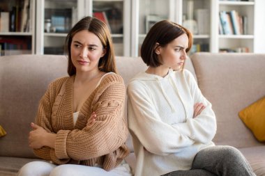 Offended angry sad pretty european millennial lady ignores girlfriend sit back to back, think about breakup in living room interior. Quarrel and relationship problems at home during covid-19 pandemic