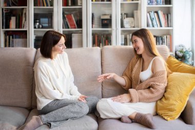 Cheerful european millennial ladies in sweaters talk, laughing, sit on sofa, enjoy meeting and free time in comfortable living room interior. Communicate together, relationships of sisters, family