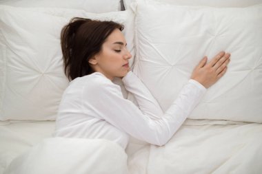 Loneliness concept. Young brunette woman lying in double bed with one hand on empty pillow next to her, miss her boyfriend, lady wearing white cotton pajamas, top view, copy space