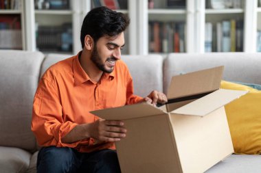 Handsome young arab man customer opening good parcel cardboard box at home satisfied with great purchase, male consumer unpacking package, happy with postal shipping delivery, copy space