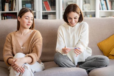 Smiling displeased european millennial lady looking at happy surprised woman with pregnancy test, rejoice positive result in living room interior. Envy, good news and baby waiting together at home