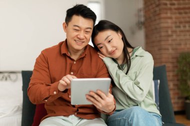Happy asian man and woman using digital tablet, watching videos or surfing internet, sitting and relaxing on couch in living room. Korean couple shopping online from home