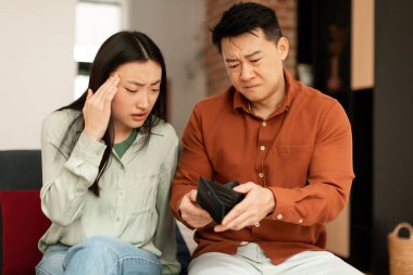 Povertry and debt concept. Unhappy asian spouses having financial problems, not able to pay bills, looking at empty wallet and searching money, sitting on couch at home