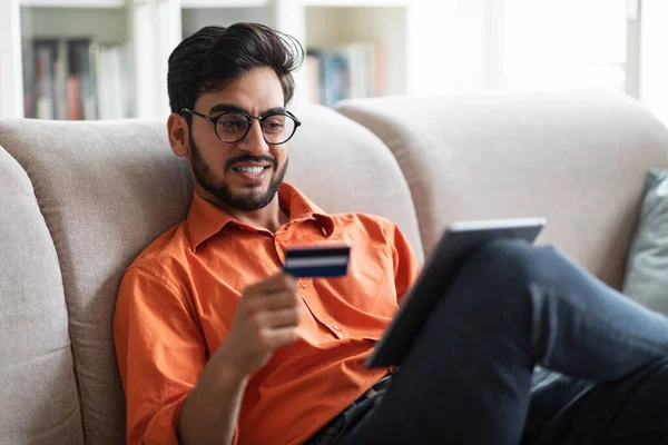 正陽気でハンサムな若いアラビア人の男が自宅でソファに座って 最新のデジタルパッドとクレジットカードを保持し 最新の銀行やショッピングモバイルアプリケーションを楽しんで コピースペース — ストック写真