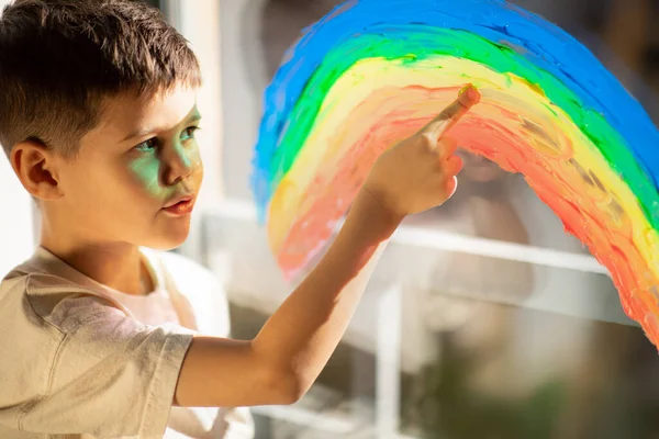 Serious little boy draws rainbow on window with hand, dream, think in room interior, sun flare, close up. Upbringing and childhood, fun and entertainment at home, art and study due covid-19 pandemic