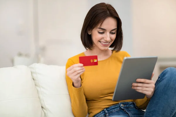 Cute cheerful pretty brunette young lady sitting on couch in living room, holding red credit card and modern digital tablet, making online order or purchasing on Internet, copy space. E-commerce