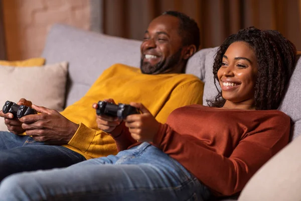 Relaxed african american spouses chilling on couch, holding joysticks and smiling, happy black couple playing video games at home, living room decorated with festive lights interior, copy space