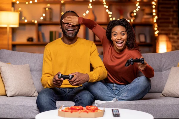 Positive funny black couple in casual outwear sitting on couch, holding joysticks, eating pizza, woman covering man eyes, cheating in game, lovers playing video games at home, copy space
