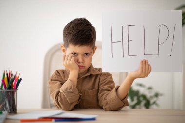 Sad unhappy little boy sits at table, studies and shows banner with inscription help in room interior, free space. Problems at education, upbringing and childhood, children whims, schooling at home