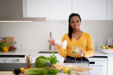 Smiling millennial african american lady in casual making smoothie with blender enjoy health care at table with organic vegetables in white kitchen interior. Weight loss with proper nutrition at home