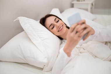 Cheerful happy beautiful young brunette woman wearing white pajamas using cell phone in bed after waking up in the morning, chatting with boyfriend, bedroom interior, copy space