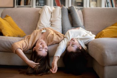 Happy european millennial ladies in sweaters lie on sofa, enjoy rest, communication and relax at free time in comfort living room interior. Relationship, fun together, gossip at bachelorette at home