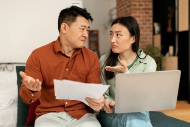 Angry korean spouses quarreling over debts and bills with papers and laptop, sitting on sofa at home. Problems with finances, bankruptcy, stress and crisis