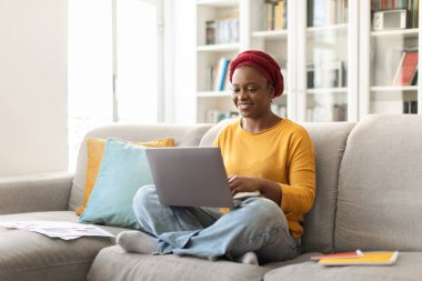 Positive pretty millennial african american woman with red turban in casual outfit sitting on couch, typing on laptop keyboard and smiling, female digital nomad working from home, copy space