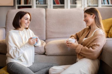 Laughing european millennial girlfriends in sweaters with coffee cups sit on sofa, enjoy meeting and talking in comfort living room interior. Gossip, good news, communication together, relationships