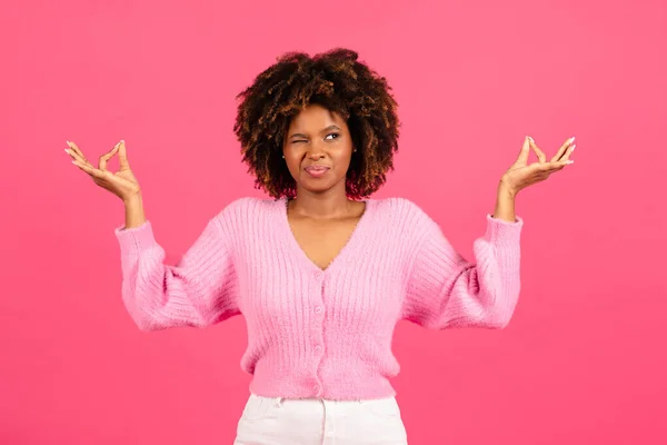 Confuse cheerful funny millennial african american curly woman in casual spreads her arms to side, looks with one eye, meditates and peeks isolated on pink background, studio. Emotions, peace, relax