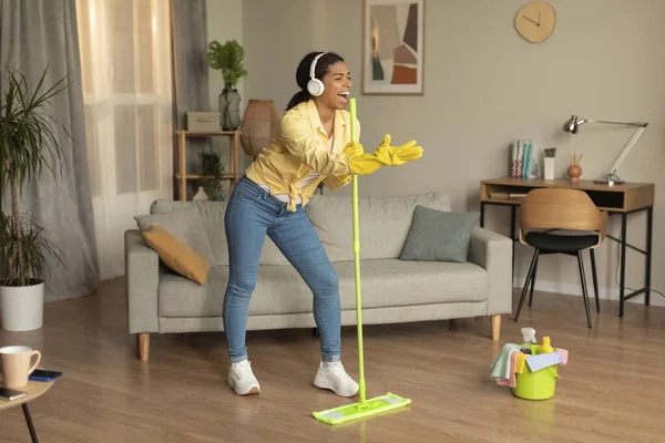 Housekeeping in fun. Excited black woman singing song while washing floor in living room, holding using mop as microphone, playful young lady gamifying cleaning routine, dancing