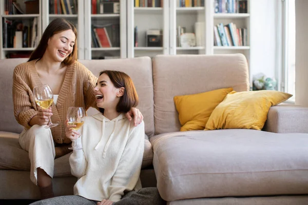 Satisfied laughing millennial european friends cheers glasses of wine, enjoy rest, gossip at spare time, celebrate holiday in comfort living room interior. Date, party, visit and fun together at home