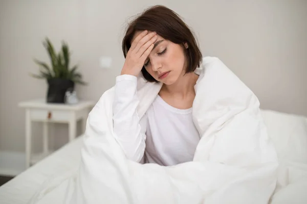 Sick unhappy young beautiful brunette woman waking up with headache in the morning, ill lady sitting in bed covered in blanket at home, touching her head, suffering from flu or coronavirus, copy space
