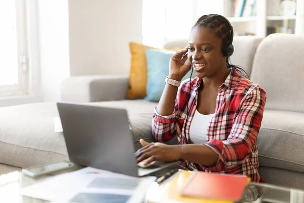 Cheerful pretty young black lady have job interview from home, sitting on floor in living room, using laptop, headset, looking at webcam screen and smiling, female teacher having online lesson
