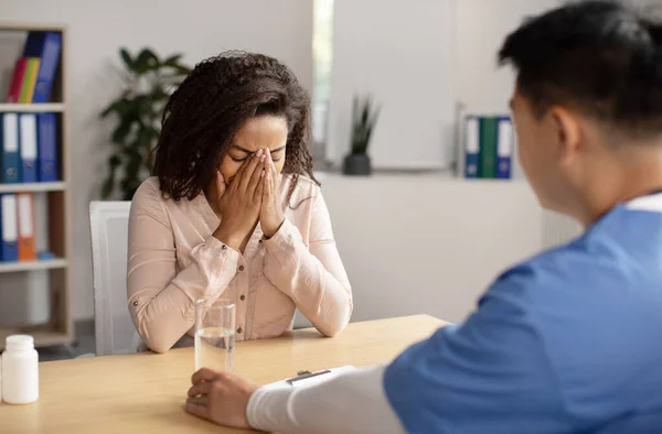 Serious middle aged korean doctor calms crying black millennial female patient, giving glass of water in clinic office interior. Health problem, visit to therapist, reaction bad news about disease